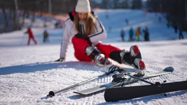 Ski on white snow with blurred slim Caucasian woman sitting at background stroking painful leg. Sad young lady fallen skiing on winter resort on sunny day outdoors