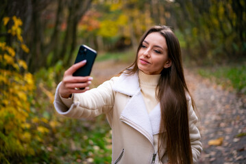 Pretty young lady making selfie in autumn park. Beautiful attractive woman with phone.