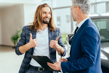 Young buyer happy with car rental terms showing thumbs up. Successful middle-aged businessman in a business suit is selling cars to a young hansome male buyer in a car showroom.