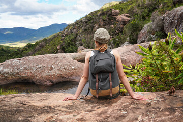 Young woman in hiking clothes, resting on stones near natural water pool, blurred trees and mountains background, view form behind - typical scenery seen during trek in Andringitra, Madagascar - obrazy, fototapety, plakaty
