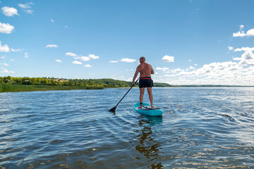 A man balances on a SUP board with a paddle in the lake on a sunny day against the backdrop of white clouds.