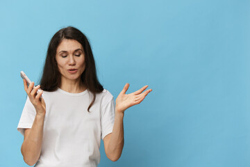 a pensive woman spreading her arms in a white T-shirt stands on a blue background with a phone in her hands with her eyes closed. Horizontal photo with space for advertising text