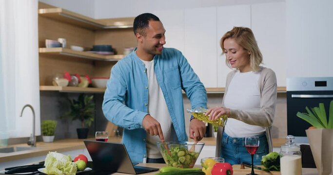 Woman pouring sunflower olive oil in white salad bowl in slow motion. Multiethnic couple in the kitchen cooking vegetable salad. Healthy Lunch. Culinary and vegetarianism.