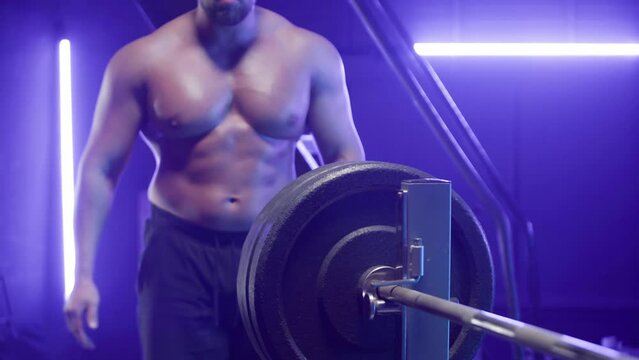 Muscular male bodybuilder with naked torso stringing round black weights onto barbell. Man with bulging muscles in gym with blue light preparing for strength training. Close up.