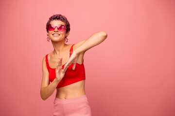 Fashion portrait of a woman with a short haircut in colored sunglasses with unusual accessories with earrings smiling on a pink bright background with a fitness body dancing