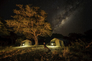 The milky way in the sky with a tents below in a bush camp as a man sits looking up at the sky; Botswana