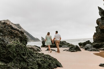 A young couple holding hands walks on a cloudy day on the beach between the rocks.