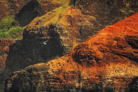 The stunning and colourful cliff sides of the Na Pali coast,  with a helicopter flying in front of them; Kauai, Hawaii, United States of America