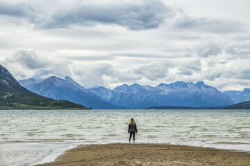 Woman stands on a sandy beach looking out at Carcross Lake and the vast Yukon Ranges; Carcross, Yukon, Canada