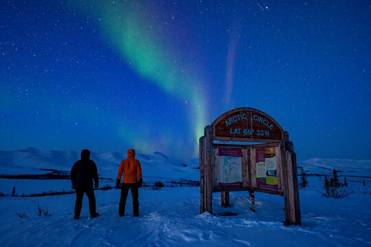 View taken from behind of two people watching the aurora borealis (or northern lights) over the Dempster Highway in the night sky at the border of the Arctic Circle; Yukon, Canada