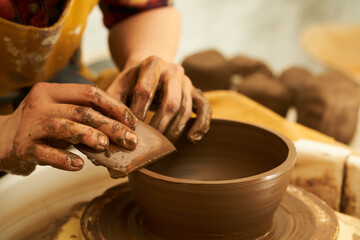 Fototapeta na wymiar A Potter works with red clay on a Potter's wheel in the workshop..Women's hands create a pot. Girl sculpts in clay pot closeup. Modeling clay close-up. Warm photo atmosphere.