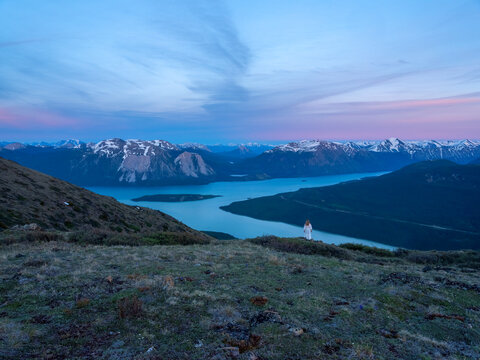 Bove Island can be seen in the centre of Tagish Lake, Yukon. Midnight sun illuminates the skies above creating a serene scene. A woman in a dress is standing overlooking the scenery.