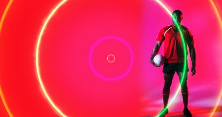 African american rugby player holding ball over colorful illuminated circles on pink background