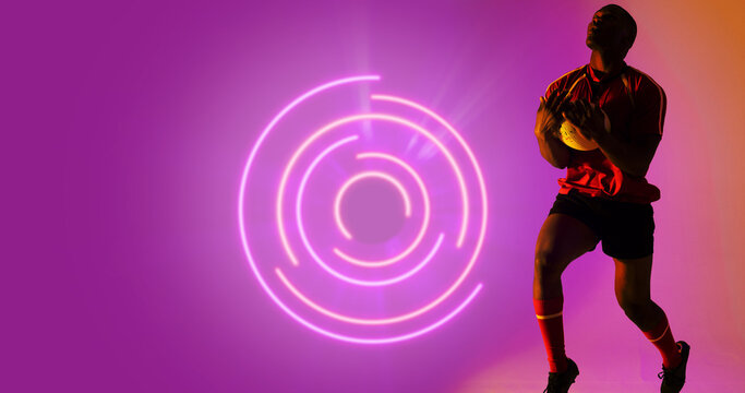 African american rugby player holding ball by illuminated circular pattern on purple background - Powered by Adobe