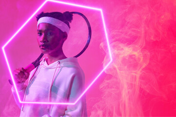 Obraz premium African american female tennis player with racket standing by illuminated hexagon on pink background