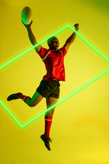 African american male player with rugby ball raising arms and jumping by illuminated rectangle