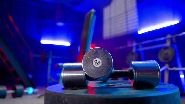 Silvery metal dumbbells lying on a blurred background of an empty gym. A rack with crossbar, barbell, weights and a sports bench in red and blue light. Sports equipment.