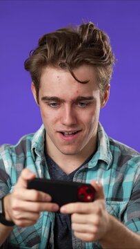 Vertical video playing game on phone smiling young man 20s in shirt play racing on mobile cell phone hold smartphone for pc video game isolated on blue background studio portrait