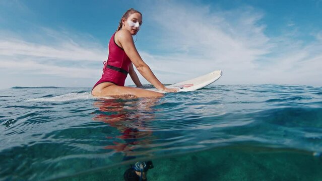 Portrait of the woman surfer in red suit with white zinc suncream on the face sitting on the surf board and mysteriously looking into the camera