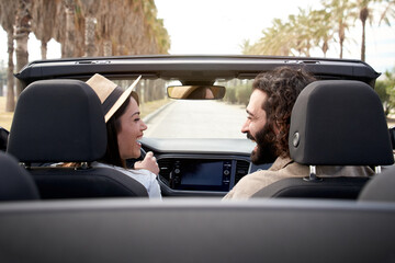 Cheerful Caucasian couple driving on sunny day in convertible car looking at each other and smiling. Touristic city road trip on a country. Urban landscape holidays happy man and woman having fun