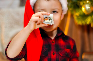 A cute boy shows Japanese rolls while sitting under the Christmas tree in a red Santa hat and a red plaid shirt.