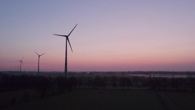 Aerial shot by a drone of windmills with rotating wings among green fields against a sunrise or sunset background. Wind farm with turbine cables for wind energy. Renewable energy source, earth care