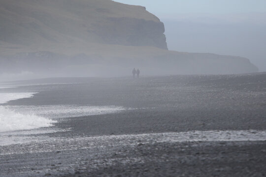 Two People Walking Away From The Camera On Reynisfjara Beach On The Southern Coast Of Iceland.