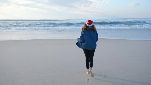 A girl or young woman in a red Santa Claus hat having fun, running and turning around on the beach Atlantic Ocean. Christmas or New Year holiday concept. 4K resolution video.