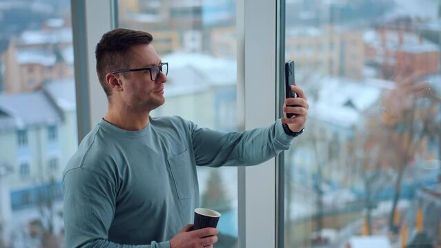 Man in glasses with a paper cup in hands standing at the window. Caucasian male having video chat on the smartphone.