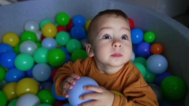 Toddler in orange shirt sitting in the soft basin among the bright balls. Baby boy tries to climb out of the pool biting the edge.