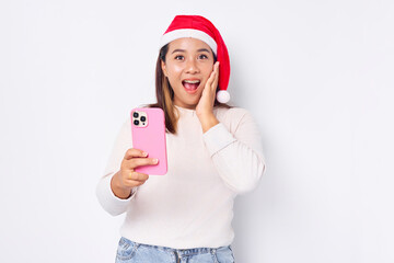 Excited young Asian woman in a Christmas hat holding mobile phone and holding cheek isolated on white background. celebration Christmas holiday and New Year concept