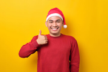 Cheerful young Asian man in Santa hat showing thumbs up, looking at camera on yellow studio background. celebration Christmas holiday and New Year concept