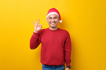 Naklejka premium Cheerful young Asian man in Santa hat showing ok sign and looking at camera over yellow studio background. celebration Christmas holiday and New Year concept