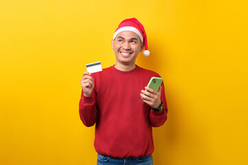 Smiling young Asian man in Santa hat using mobile phone and credit card, looking up, over yellow...