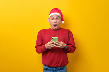 Surprised young Asian man in Santa hat holding smartphone, looking at camera with opened mouth over yellow studio background. celebration Christmas holiday and New Year concept