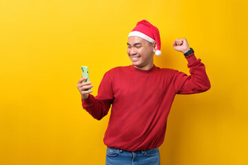 Fototapeta na wymiar Excited young Asian man in Santa hat using smartphone and doing winner gesture over yellow studio background. celebration Christmas holiday and New Year concept