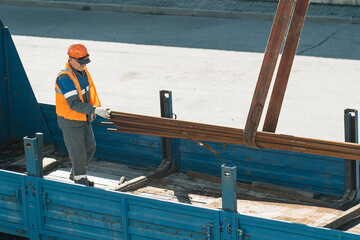 Naklejka premium Slinger in helmet and vest unloads metal pipes and fittings from truck body on clear day.. Production background. Authentic workflow on construction site. Slinger at work..