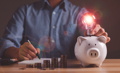 Businessman holding light bulb on piggy bank and writing notebook, creativity, financial accounting, saving energy and money concept.