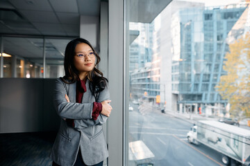 Asian businesswoman with arms crossed looking through window in office. - Powered by Adobe