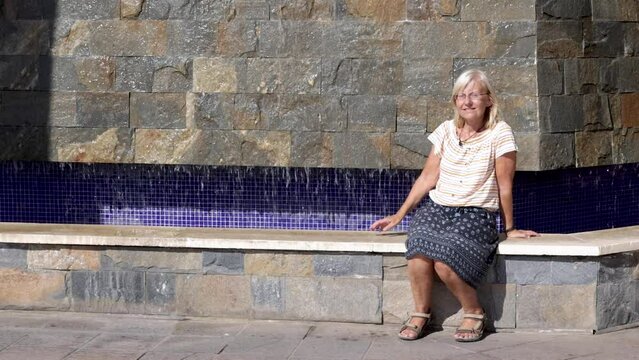 Blond woman sitting and enjoying the sun while looking around by a small artificial waterfall outside La Zenia shopping center in Torrevieja, Spain
