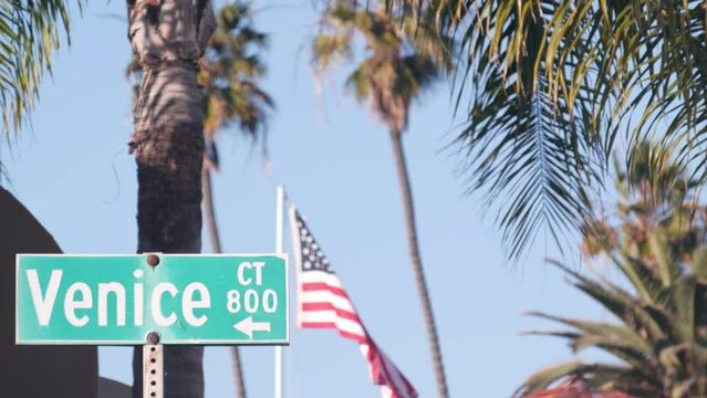 Venice beach road sign on crossroad, California city street, USA. Waterfront tourist resort in Los Angeles, beachfront travel destination, waterside summer vacations. Coastal palm trees. American flag