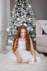 A cute girl with long wavy hair in a white dress on the background of a Christmas tree with a rabbit in her hands.