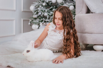 A cute girl with long wavy hair in a white dress on the background of a Christmas tree with a rabbit in her hands.