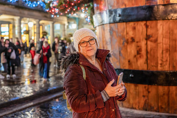 Happy senior woman tourist in a knitted hat, jacket, with a backpack walks along the street in a rainy festive city, smiling, and used phone. Christmas lights lights and crowd in the background.