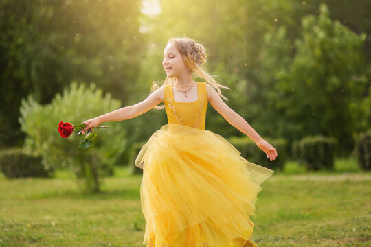 A little girl with blond hair in a lush yellow dress walks in the park with a red rose. Princess image.