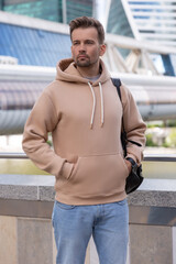 City portrait of handsome hipster guy with beard wearing beige blank hoodie or sweatshirt with space for your logo or design. Holds a backpack over his shoulder. Mockup for print
