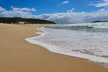 Waves crashing at Fingal Beach with people walking along the sand in the distance on a sunny day.