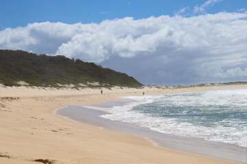 Waves crashing at Fingal Beach with people walking along the sand in the distance on a sunny day.