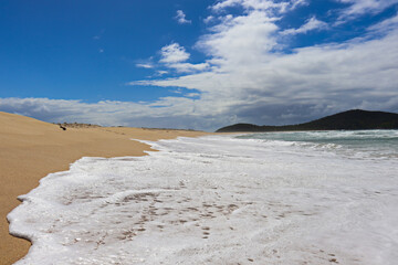 Waves crashing at Fingal Beach with people walking along the sand in the distance on a sunny day.