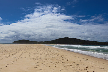Waves crashing at Fingal Beach with people walking along the sand in the distance on a sunny day.